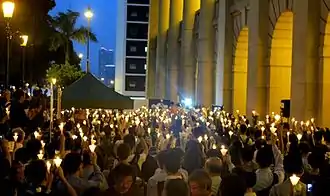 Quelques centaines des  mille cinq cent manifestants chinois présents pour le septième jour du décès de Li Wangyang, massés au premier plan à droite devant les arcades éclairées du Conseil législatif à Hong Kong, et tenant des cônes de papier enserrant une bougie. Buildings, palmiers, lampadaires  et ciel bleu-nuit éclairée en arrière-plan.