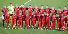 Photographie de face de onze joueurs de football au maillot rouge, derrière onze enfants.