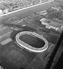 Photographie en noir et blanc. Vue aérienne d'un stade avec des tribunes, près d'un autre terrain sans tribunes et d'un cours d'eau.