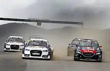 Cinq voitures de sport vues de face sur une piste goudronnée et soulevant un nuage de poussière.