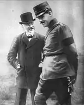 photographie noir et blanc : deux hommes debout ; au fond un homme barbu en chapeau ; devant un homme moustachu en uniforme