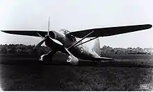 Photo en noir et blanc d'un avion à hélice posé sur une surface en herbe, à l'arrière-plan des habitations.