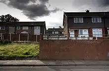 Depuis une rue, vue sur des maisons britanniques du XXe siècle. Les maisons sont surélevées par rapport à la rue. Un terrain vague et herbeux apparaît à la gauche et une mur de briques se trouve à la droite.