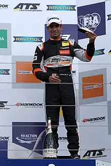 Photographie d'un pilote indien, brun en combinaison noire, vu de face, debout sur un podium, brandissant un trophée dans sa main gauche, souriant.