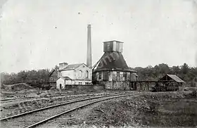 Photo en noir et blanc de petits bâtiments, avec grande cheminée et tour pyramidale en bois fermée en forêt.