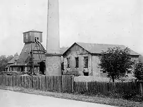 Photo en noir et blanc de petits bâtiments, avec grande cheminée et tour pyramidale en bois fermée.