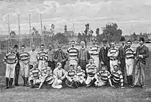 Un groupe d'hommes, habillés en majorité d'un polo sportif, pose en rang pour une photographie. En arrière-plan, des installations sportives.