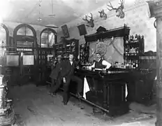 la photo noir et blanc représente deux hommes accoudés à un bar en bois derrière lequel le patron prend la pose.