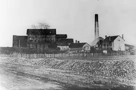 Photo noir et blanc montrant à gauche un très grand bâtiment en bois entouré de plusieurs autres en bois ou maçonnés accompagnés d'une cheminée à base carrée sur la droite.