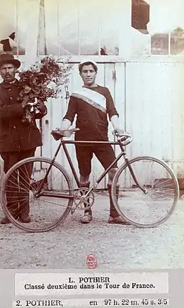 Photographie en noir et blanc d'un homme tenant son vélo et portant une casquette, une tenue sombre et une écharpe en bandoulière.
