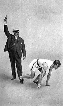 Photographie en noir et blanc de deux hommes. Celui de gauche est en costume, porte un chapeau, pistolet à la main. Celui de droite est en position de départ d'une course