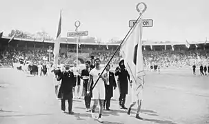 Une photo noir et blanc de juillet 1912. Sur la piste d'athlétisme du stade olympique de Stockholm, s'avance, la délégation japonaise, composée de deux sportifs en short et maillot blanc et quatre officiels en costume sombre. L'un des sportif, au tout premier plan, porte un drapeau japonais.