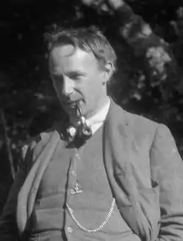 Photographie en noir et blanc d'un homme élégamment vêtu au regard pénétrant, fumant la pipe, une chaîne de montre en évidence sur le gilet de son costume.