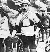 Photographie en noir et blanc d'un coureur cycliste se tenant debout à côté de son vélo au départ d'une course.
