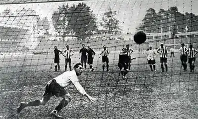 Photographie en noir et blanc, vue d'un but de football : le gardien n'arrive pas à toucher le ballon, tiré par penalty