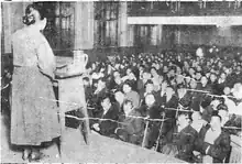 Photographie d'un meeting politique. Au premier plan une femme de dos s'adresse au public, visible dans le fond de l'image, constitué de rangs de personnes assises.