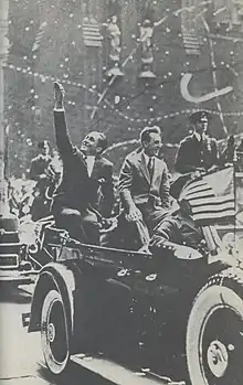 Photographie en noir et blanc montrant deux hommes saluant la foule, assis à l'arrière d'un véhicule.