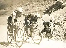 Photographie en noir et blanc de deux cyclistes montant un col, l'un poussant l'autre, un homme courant à leur côté.