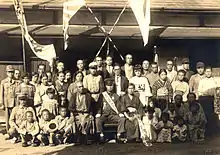 Une famille se rassemble autour d'un garçon en uniforme, entourée de bannières et drapeaux. Des enfants en ont aussi.