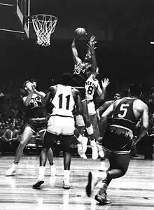 Image en noir blanc de Wilt Chamberlain sautant pour attraper le ballon en l'air au-dessus d'autres joueurs, à proximité d'un panier de basket-ball.