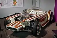 A white car with brown stripes, with open wheels and a clear bubble canopy over twin seats, and exposed, chromed engine with a blower.  Beatnik Bandit II and a few of Roth's other cars are also on display in this museum.