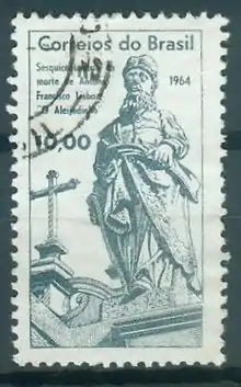 Photographie en couleur d'un timbre postal sur un support noir. L'encre est verte et le motif représente une statue en premier plan, devant le toit de l'église du sanctuaire.