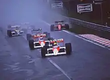 Photo de plusieurs monoplaces en formation rapprochée. Senna est en tête suivi de Prost (F1 rouges et blanches), Berger, Boutsen et Mansell (avec deux roues dans l'herbe).