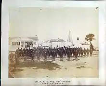 Union engineers lined up in sandy foreground of white houses, white lighthouse in the far background