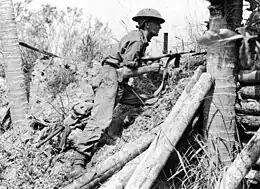 Photo en noir et blanc d'un soldat tenant une mitrailleuse, allongé contre un talus dans la jungle.
