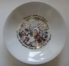 Photo d'une assiette décorée commémorative de la félibrée de 2001 à Périgueux. Le dessin représente un couple de périgourdins en costume traditionnel en tête d'un défilé. La femme sur la droite joue du violon et l'homme à gauche de l'accordéon.
