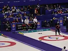 Photographie d'une piste de curling sur laquelle se trouve deux curleurs portant un balai, devant des gradins.
