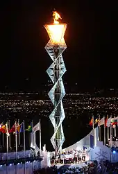 Photographie d'une structure métallique surplombant un stade, au sommet de laquelle brûle une flamme, la nuit. Des hommes sont rassemblés à son pied.