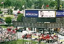 Phtographie en couleur d'un paddock de course, avec des spectateurs à gauche, et un tableau des scores au centre, surmonté de plusieurs drapeaux.