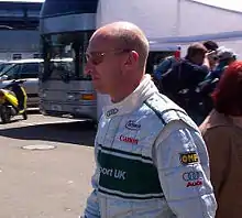 Photo de profil gauche d'un homme en combinaison blanche et verte, portant des lunettes de soleil, devant un camion