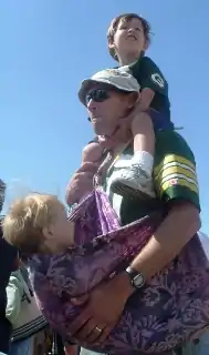 Homme portant un enfant sur ses épaules et un autres en écharpe.