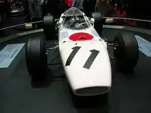 Photographie en couleur de la Honda RA272 blanche avec un cercle rouge, vue de face, dans un musée d'exposition.