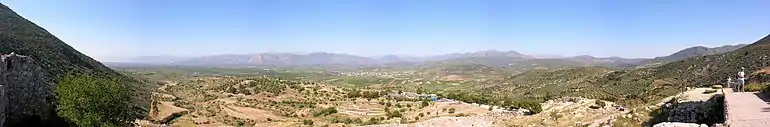 Panorama d'une plaine, prise depuis une hauteur. Aux extrémités gauche et droite, des flancs de collines. Au fond, après la plaine, une chaîne de montagnes basses. Au premier plan, les ruines de la cité de Mycènes.