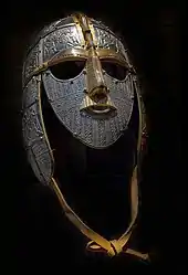 Photographie d'un casque métallique équipé de garde-joues, d'un masque facial et d'une jugulaire en cuir. La surface métallique est décorée avec des motifs finement ciselés, tandis que certains éléments sont dorés.