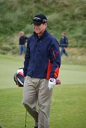 Joueur de golf debout, couvert d'une casquette, et vetu d'un blouson bleu.