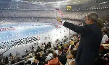 Homme debout dans une tribune d'un stade, le bras gauche avec un drapeau américain tendu pour saluer des athlètes.