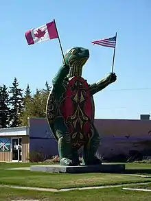 Une grande statue représentation une tortue en position bipède, tenant un drapeau canadien dans une main et un drapeau américain dans l'autre.