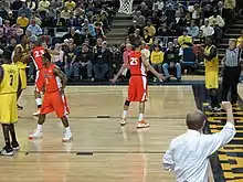 Remise en jeu lors d'un match de basket-ball par un joueur sur la droite. En bas à droite, un homme de dos, en chemise, lède un bras.