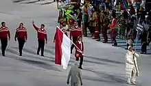 vue partielle d'un défilé : une femme en grande doudoune blanche brandi une pancarte marquée « MONACO », suivie à quelques mètres d'une femme en veste 3/4 de lainage rouge, décorée de quelques losanges blancs, portant un drapeau rouge et blanc en deux bandes horizontales égales ; suivent 5 hommes de front, en pantalon sombre et pull-over rouge ornés de losanges blancs aux épaules, dont l'un salue la foule du bras.