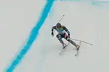 Photographie d'un skieur dans la descente, jambes fléchies, portant une combinaison grise et noire et le dossard numéro 16.