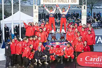 Sébastien Loeb et Daniel Elena debout sur le toit de leur Citroën C4 WRC, levant les bras au ciel et entourés de l'équipe de Citroën Racing.