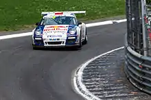 Porsche 997 GT3 Cup de couleur bleue et blanche, pilotée par Sébastien Loeb et vue de face dans un virage sur une piste goudronnée.