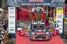 Sébastien Loeb et Daniel Elena sabrant le champagne debout sur le toit de leur Citroën DS3 WRC, portières ouvertes et entourée de spectateurs.
