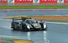 Photographie d'une voiture de sport-prototype, vue de trois-quarts, noire et or, sur une piste humide.