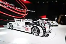 La 919 Hybrid et la 919 RSR exposées sur un stand du salon avec une robe blanche et grise, un écran géant est visible derrière elles.