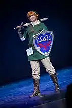 Jeune homme blond avec une tunique verte, portant une épée.